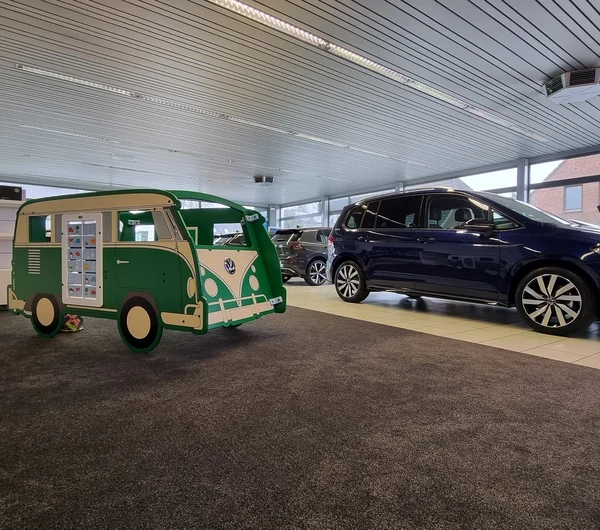Groene IKC speelbus in de stijl van een klassieke Volkswagen, geplaatst op tapijt in een lichte showroom. Op de achtergrond staan diverse auto's opgesteld; links op de vloer liggen een strandbal en speelgoed.
