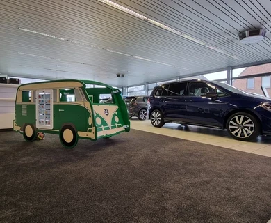 Groene IKC speelbus in de stijl van een klassieke Volkswagen, geplaatst op tapijt in een lichte showroom. Op de achtergrond staan diverse auto's opgesteld; links op de vloer liggen een strandbal en speelgoed.