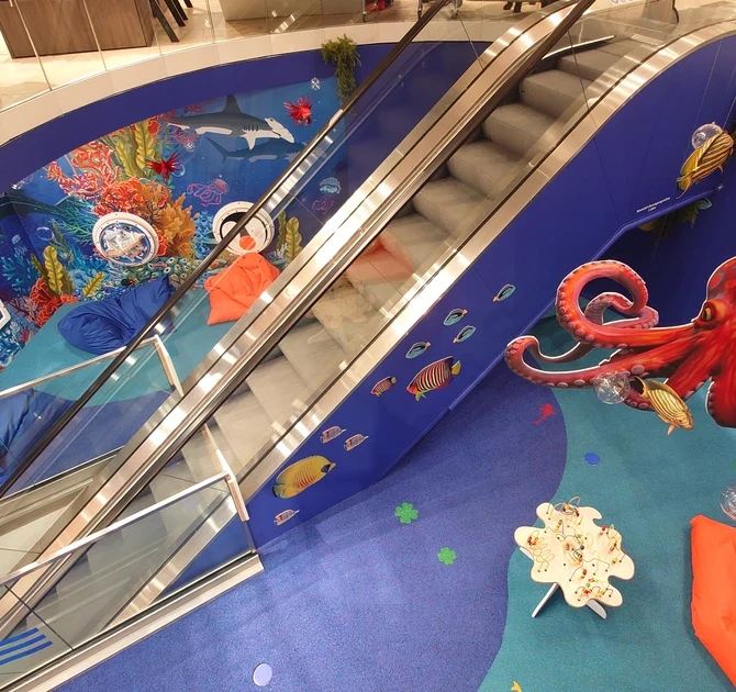 Kinderhoek in oceaanthema rondom een roltrap, met wandspellen, kleurrijke onderwaterillustraties, grote octopusdecoratie en zachte zitzakken, ontworpen door IKC in een winkelcentrum.