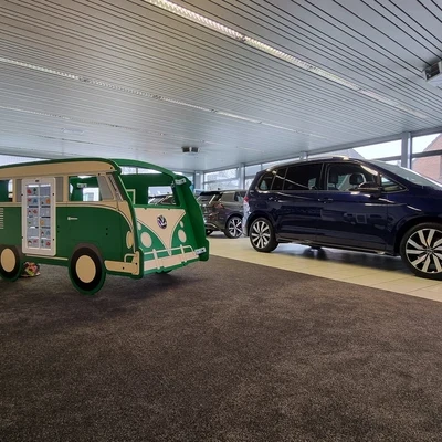 Groene IKC speelbus in Volkswagenstijl met wandspel, geplaatst op een donkergrijs tapijt in een moderne showroom. Rechts in beeld meerdere auto’s, links een strandbal en kinderspeelgoed.